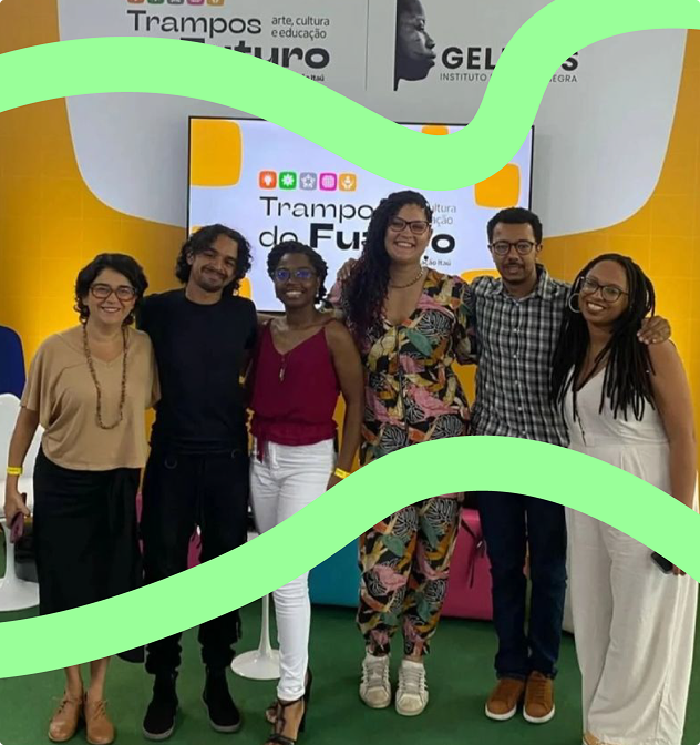 Aparecem, da esquerda para a direita, as seguintes pessoas: Fernanda Nobre, Frances Gonf, Lassalette Jorge, Ariely Tombô, Amauri Eugênio Jr. e Natália Carneiro. Elas estão abraçadas e à frente do palco, que é composto por uma parede laranja com detalhes brancos, dentro dos quais aparecem os logos do evento Trampos do Futuro e do Geledés - Instituto da Mulher Negra, assim como uma smart TV. Há, ao fundo, pufes coloridos nos quais as pessoas participantes estiveram sentadas durante a roda de diálogo.