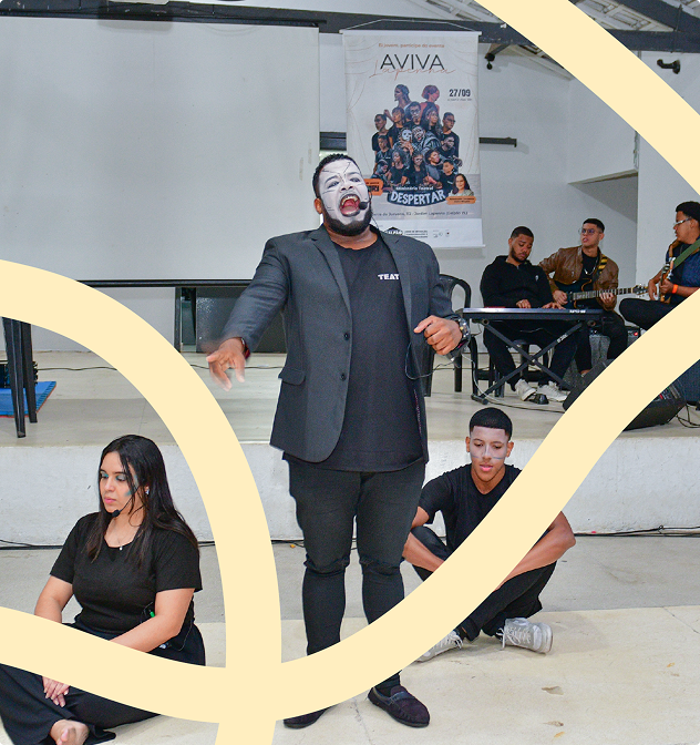 Intervenção teatral durante o Aviva Lapenna. Há um homem ao centro que está com maquiagem na cor branca e usa camiseta e blazer pretos - ele aparece cantando. Além dele, outras pessoas aparecem ao redor dele e sentadas no chão.