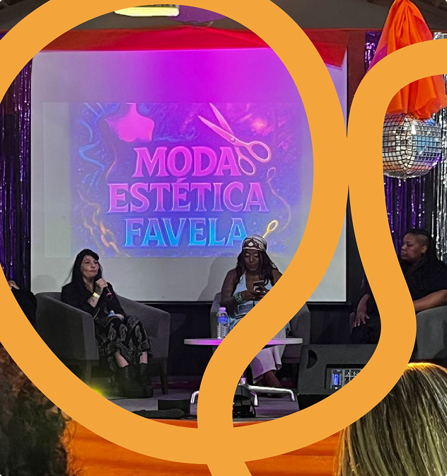 Imagem de cinco pessoas no palco do Galpão ZL. As três pessoas mais ao centro estão sentadas, cada uma, em uma poltrona. Os dois homens nas extremidades esquerda e direita estão sentados em cadeiras. Pode-se ver também, ao centro, uma caixa de som. As pessoas estão à frente de uma tela na qual é possível ler o título da edição do Festival Cria na Vila (Moda Estética Favela) nas cores azul e rosa, sobre um fundo escuro, assim como os desenhos de uma tesoura e de um manequim. Há, também, fitas coloridas decorando o palco, assim como globos brilhantes instalados no mesmo espaço.