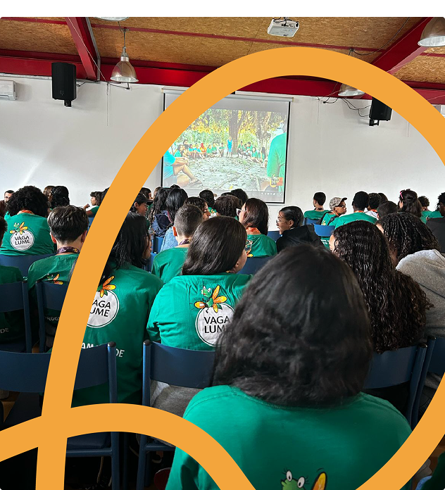 Imagem de reunião da Rede Vaga Lume. As crianças e adolescentes participantes aparecem de costas, com camisetas do projeto, sentadas em cadeiras dispostas em um salão.