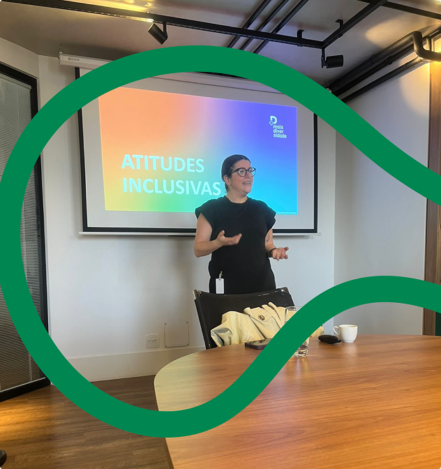 Imagem da pessoa responsável pela condução da atividade sobre atitudes inclusivas no ambiente profissional. Ela está em pé, à frente de uma tela que apresenta um slide com nas cores lilás, azul-claro e laranja, que contém a mensagem Atitudes inclusivas. Ela está atrás de uma cadeira e de uma mesa.