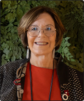 Imagem de Maria Alice Setubal. Ela tem cabelos castanhos, com corte chanel, na altura do pescoço, usa óculos, xale preto com detalhes pontilhados brancos e uma blusa vermelha. Ela está à frente de fundo composto por folhas.