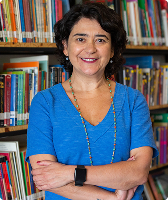 Imagem de Fernanda Nobre. Ela tem cabelos na altura do pescoço e aparece sorrindo na foto. Fernanda usa um conjunto azul-claro, colar, está com os braços cruzados e aparece à frente de uma estante com livros.