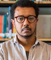 Imagem de Amauri Alfredo Eugênio Júnior. Ele usa camisa com tonalidade ocre, camiseta verde e óculos de acetato redondos, tem cabelos crespos em formato black power e está de braços cruzados. Amauri aparece à frente de uma estante com livros.