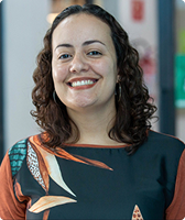 Imagem de Joyce Almeida Costa. Ela tem cabelos na altura do ombro, usa par de brincos em formato de argola e camiseta com predominância da cor verde, detalhes em laranja e estampa florida. Joyce está à frente da entrada do espaço coworking do Galpão ZL.