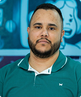 Imagem de Carlos Rodrigues Marques. Ele tem cabelo curto e barba, e usa uma camiseta polo verde. Ele está à frente de uma parede azul com desenhos na cor azul-clara.