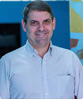 Imagem de Celso Felipe da Silveira. Ele tem cabelo curto e usa camisa na cor branca. Celso está à frente de uma parede azul com detalhes em amarelo, branco e vermelho.