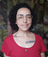 Imagem de Lorena Pereira Assis. Ela tem cabelo cacheado na altura dos ombros e usa óculos de aro fino. Lorena está com uma camiseta vermelha está à frente de uma parede colorida com estampas florais.