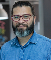 Imagem de Marcelo Ribeiro Silva. Ele aparece com cabelos presos, barba, óculos de acetato com tonalidade grená e camisa jeans. Marcelo está à frente de fundo composto por paredes pretas e brancas.