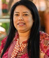 Imagem de Antônia Marlucia Martins Gomes. Ela tem cabelos longos e usa uma camisa com estampas nas cores rosa, amarelo, laranja e marrom. Malu aparece à frente de fundo composto por parede laranja, plantas e iluminação solar.