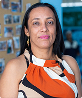 Imagem de Vânia Silva. Ela tem cabelos longos e usa blusa regata com estampa nas cores laranja, branca, rosa-claro e preto. Vânia está à frente de um mural de madeira para fotos e de paredes na cor azul.