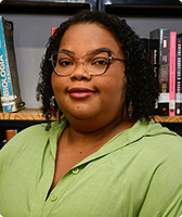 Imagem de Mauricélia Martins Catão na qual ela aparece à frente de uma prateleira com livros. Mauricélia veste uma blusa na cor verde e usa óculos e tem cabelo encaracolado na altura do pescoço.