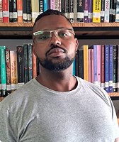 Imagem de Abraão Ferreira da Silva. Ele aparece à frente de uma prateleira com livros, usa uma camiseta cinza e óculos. Abraão tem barba e é um homem negro.