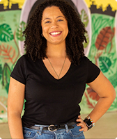 Imagem de Kelly Cristina dos Santos Juremeira. Ela tem cabelos cacheados na altura do ombro e aparece sorrindo. Kelly usa uma camiseta na cor preta e calça jeans, e aparece à frente de um mural com desenhos compostos por folhas, horizonte e céu que emula um pôr do Sol.
