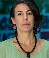 Imagem de Fabiana Tock. Ela tem cabelo encaracolado na altura do ombro, usa par de brincos metálicos, colar na cor preta e uma camiseta verde. Fabiana está à frente de parede azul na qual há desenhos nas cores verde, azul e roxa.