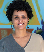 Imagem de Viviane Soranso dos Santos. Ela tem cabelo black power e cacheado e usa um par de brincos de argola. Viviane está com uma camiseta nas cores cinza e branca e aparece à frente de uma parede azul com desenhos nas cores amarela e vermelha.