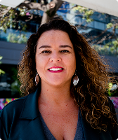 Imagem de Marília Câmara de Assis. Marília aparece com cabelos cacheados com tonalidade castanha, usa par de brincos em formato losangular, blusa azul e blazer na mesma cor.