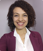 Imagem de Kátia Ramalho Gomes. Ela tem cabelos encaracolados. Kátia usa um par de brincos de argola, camisa branca e blazer roxo, e está à frente de uma parede lilás. A imagem mostra, ao fundo, a vista externa da rua Leôncio de Carvalho, na região da avenida Paulista.
