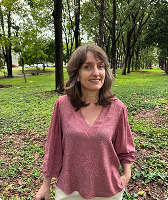 Imagem de Analine Almeida Specht. Ela tem cabelos na altura dos ombros. Analine usa uma blusa cor-de-rosa e está à frente de fundo composto por árvores.