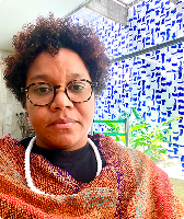 Imagem de Kelly Regina Santos da Silva. Kelly tem cabelos curtos cacheados e usa óculos de acetato. Ela usa uma camiseta preta, blusa marrom e colar branco, e está à frente de parede composta por azulejos brancos com detalhes azuis.