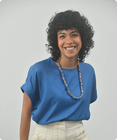 Imagem de Raissa Gonçalves Monteiro. Raíssa tem cabelos cacheados na altura dos ombros, usa camiseta na cor branca, colar e calça branca. Raíssa está à frente de uma parede branca.