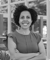 Imagem na qual Roseli Faria aparece em primeiro plano. Ela tem cabelo crespo em formato black power, usa uma blusa azul, está de braços cruzados e sorrindo. Há, ao fundo, a fachada de um edifício com vidros espelhados, nos quais estão refletidos árvores e arbustos.