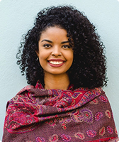 Trata-se da foto de Lígia Batista. Ela tem cabelos encaracolados e usa uma echarpe com tons de magenta. Ela aparece sorrindo na imagem.