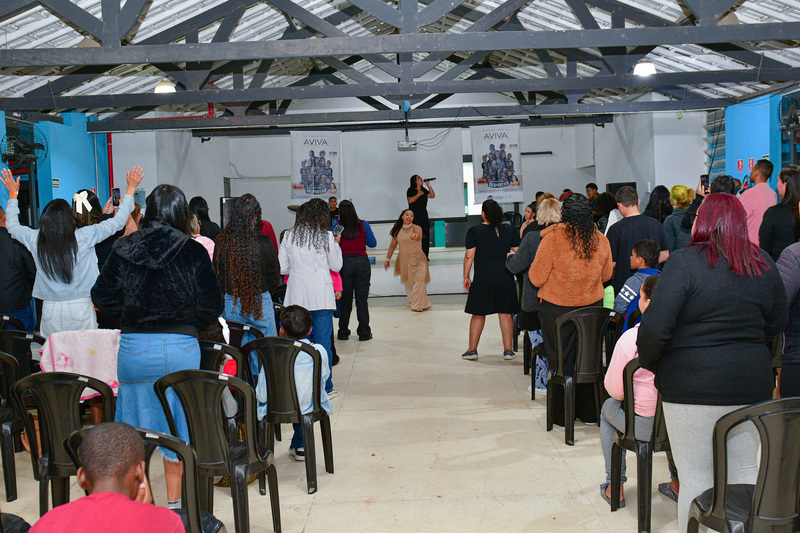 Momento de celebração religiosa no Aviva Lapenna no Galpão ZL, com pessoas em oração e uma mulher conduzindo a pregação no palco.