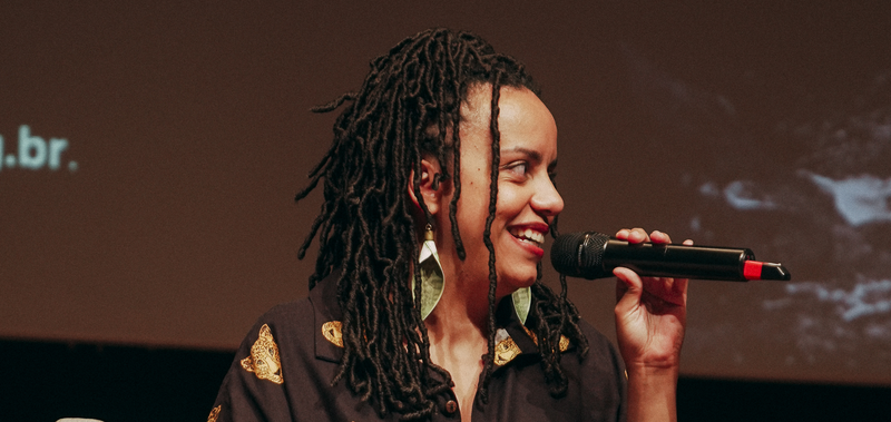 Ana Sanches falando ao microfone durante evento, com cabelos em dreads finos e brincos em formato de concha.