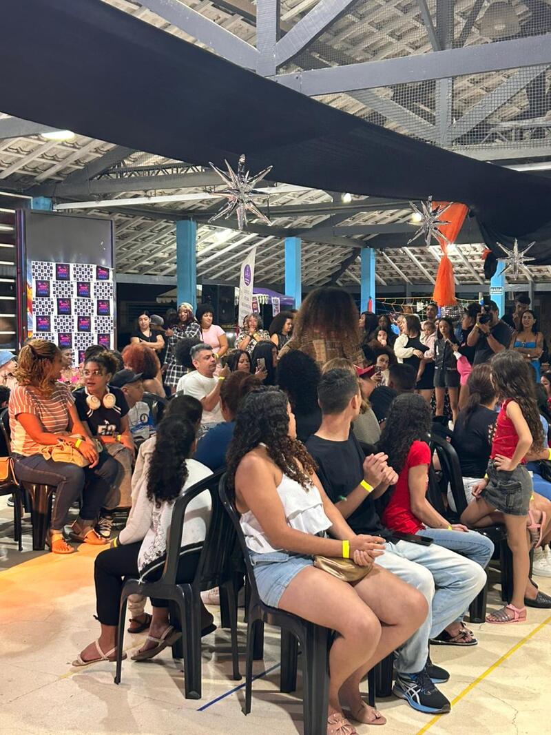 Pode-se ver pessoas sentadas em cadeiras instaladas no auditório do Galpão ZL, enquanto outras aparecem em pé - mais ao fundo.