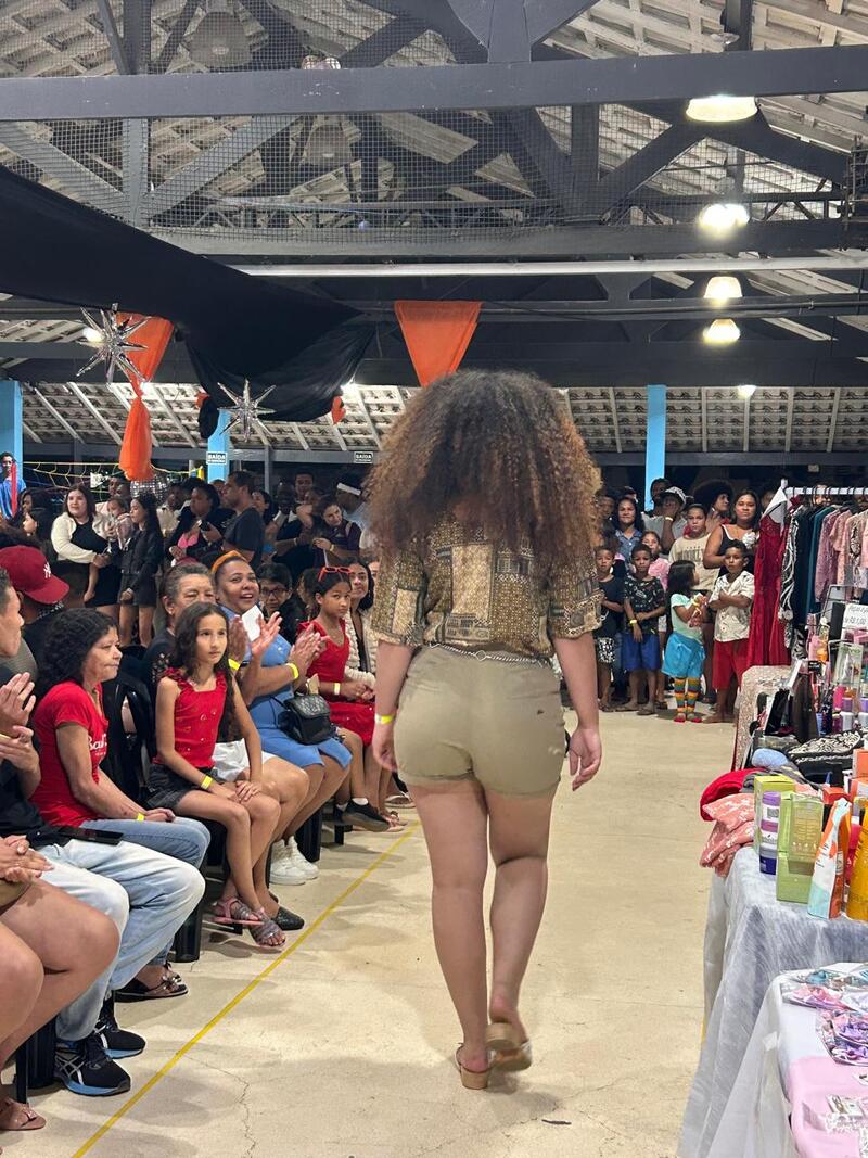 Pode-se ver pessoas sentadas em cadeiras instaladas no auditório do Galpão ZL, enquanto outras aparecem em pé - mais ao fundo. Aparece ao centro da imagem uma pessoa desfilando em uma passarela projetada para o evento.