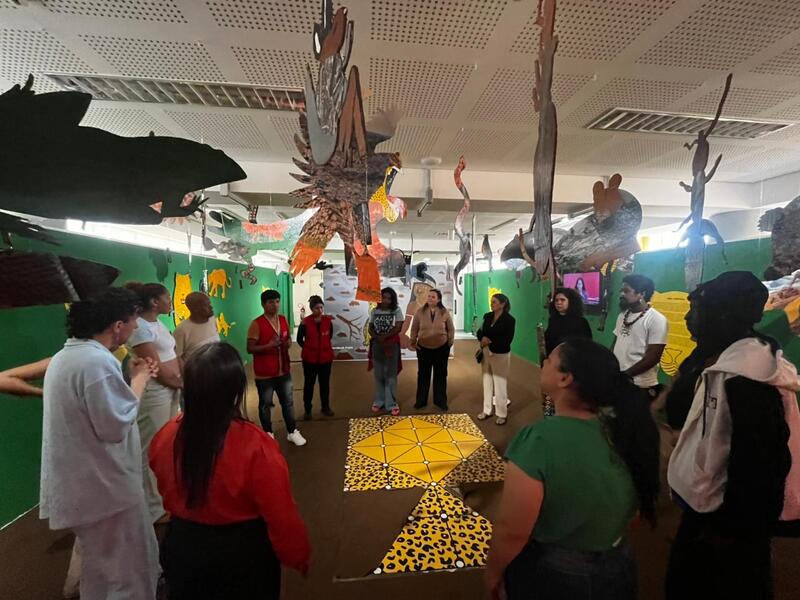 Equipe do projeto Primeiras Embarcações em roda durante visita ao Museu da Cultura Indígena, ouvindo orientações com artesanatos suspensos ao fundo.