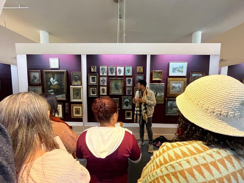 Equipe do projeto Primeiras Embarcações durante visita ao Museu Afro Brasil Emanoel Araujo ouvindo orientações, com instalação de paredes roxas ao centro.