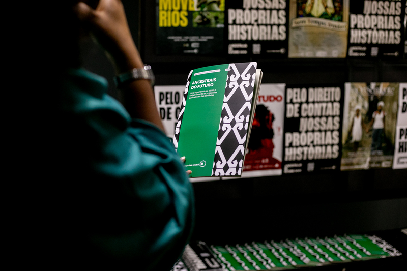 Pessoa segurando publicação da websérie Ancestrais do Futuro diante de parede com cartazes do projeto.