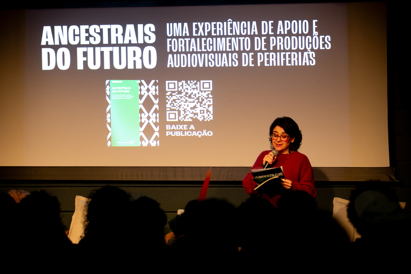 Fernanda Nobre falando ao microfone com material da websérie Ancestrais do Futuro em mãos diante de tela de apresentação.