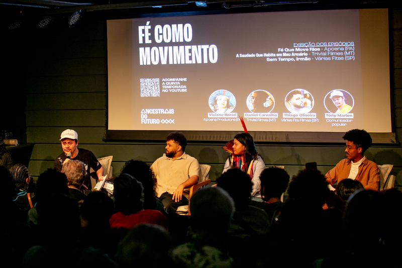 Participantes sentados no palco durante evento com plateia ao fundo.