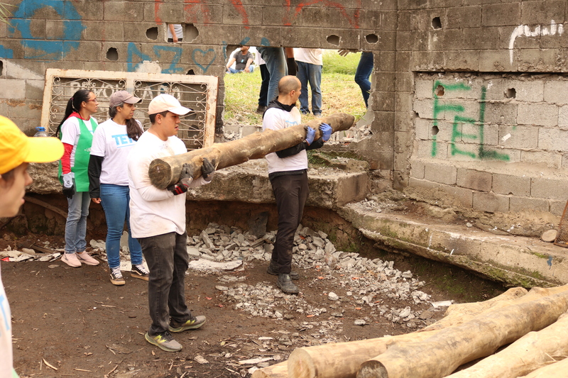 Imagem de duas pessoas carregando uma viga usada na sustentação de uma das casas do projeto +Lapena Habitar. Há outras perto deles - e esse grupo aparece à frente de um muro.