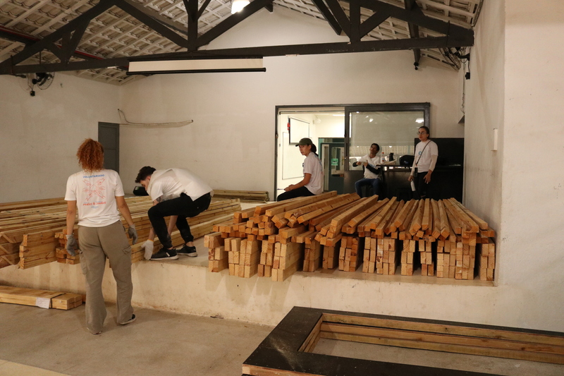 Imagem de um grupo de cinco pessoas pegando pedaços de madeira, acomodados dentro do Galpão ZL, usados na construção das casas do projeto +Lapena Habitar.