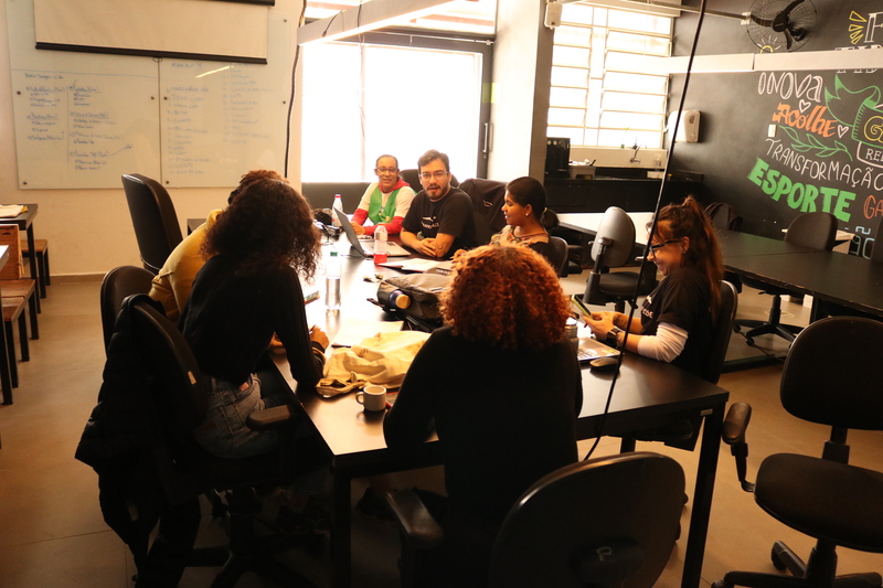 Imagem de pessoas sentadas em frente a uma das mesas do coworking do Galpão ZL. Elas conversam sobre o plano de ação para a construção das casas do projeto +Lapena Habitar.
