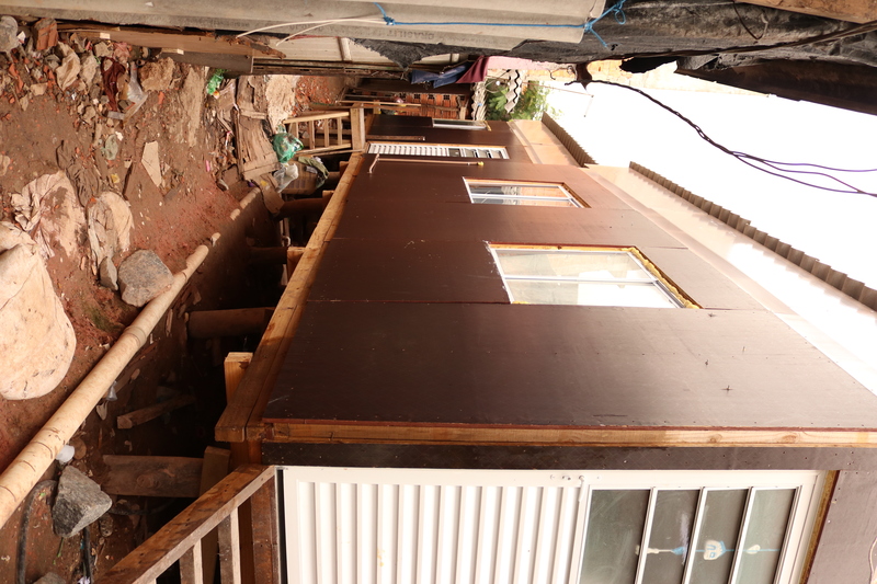 Casas do projeto +Lapena Habitar sobre palafitas em solo úmido, com pedras e encanamentos visíveis, estruturas com placas pretas.