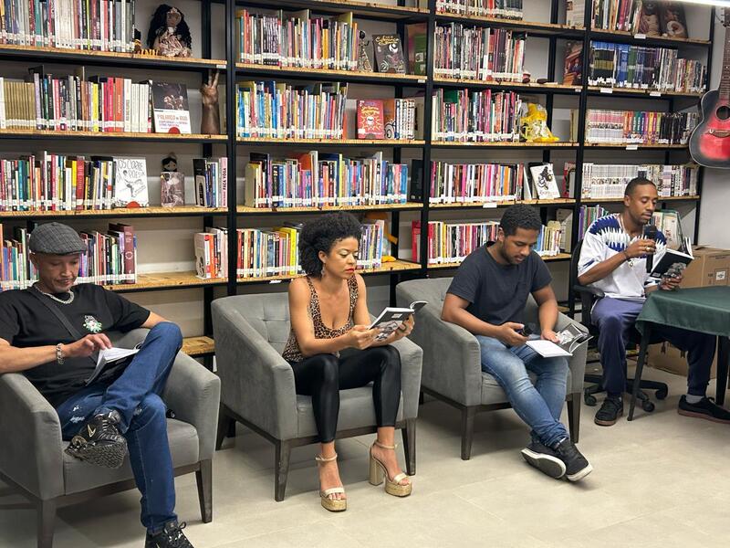 Imagem do lançamento do livro A Caminhada, de Andrio Candido. A foto mostra três pessoas sentadas em poltronas - cada uma lendo um exemplar da obra -, enquanto Andrio está com camiseta branca e calça jeans, sentado em frente a uma mesa, lendo o livro - ele usa um microfone para se comunicar com o público. Todas as pessoas estão à frente de uma estante com outras obras.