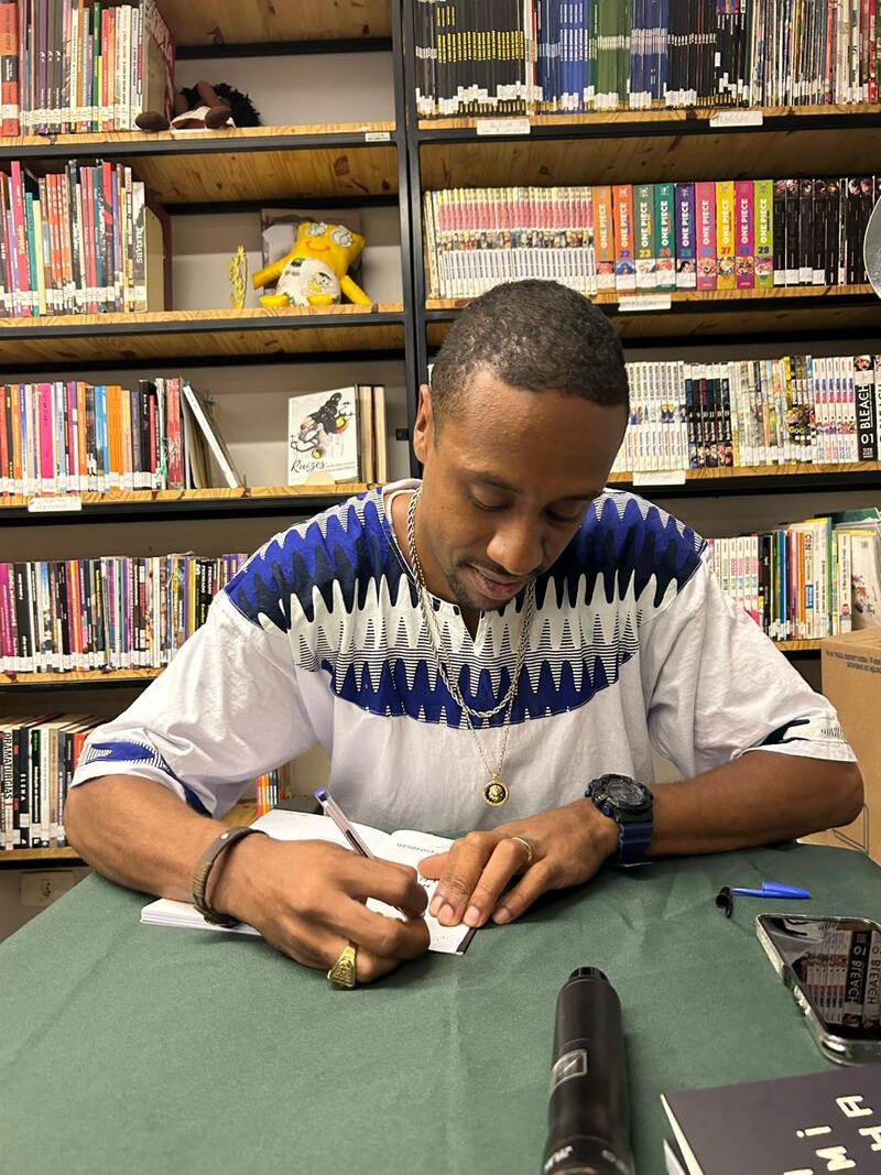 Imagem de Andrio Candido autografando um livro. Ele é um homem negro e tem cabelo crespo raspado - e usa uma camiseta branca. Ele está à frente de uma estante com livros.