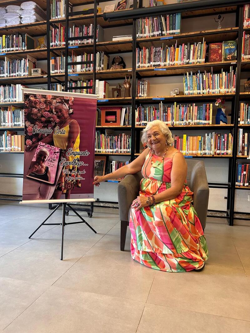 Foto de Conceição Costa, autora do livro Enquanto Envelheço. Conceição está sentada em uma poltrona e usa um vestido colorido com detalhes em bege, rosa, laranja e verde - e está ao lado de um banner de divulgação da obra. Ela está à frente de uma estante com livros.