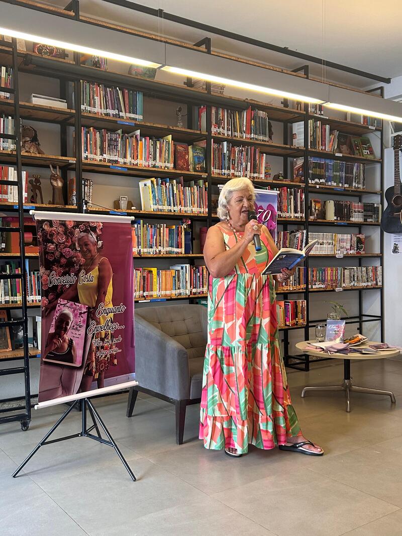 Foto de Conceição Costa, autora do livro Enquanto Envelheço. Conceição está em pé e usa um vestido colorido com detalhes em bege, rosa, laranja e verde - e está ao lado de um banner de divulgação da obra. Ela está à frente de uma estante com livros enquanto lê a obra e uma um microfone para fazê-lo.