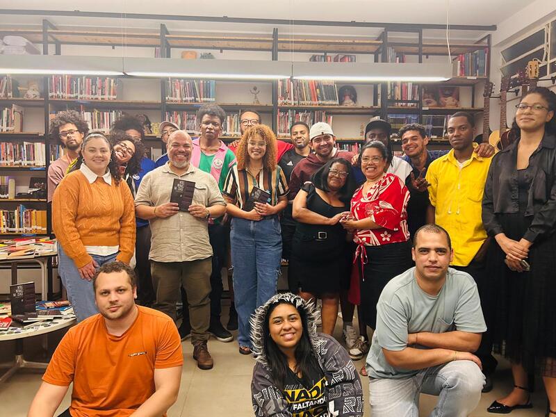 Foto do lançamento do livro Notas Infames da Cidade, de Ricardo da Paz. A imagem mostra 20 pessoas na imagem, sendo que algumas seguram exemplares da obra. Uma dessas pessoas é o autor - ele é um homem careca, com barba e camisa manga curta na cor bege, e aparece sorrindo. Todas as pessoas estão à frente de uma estante com livros.