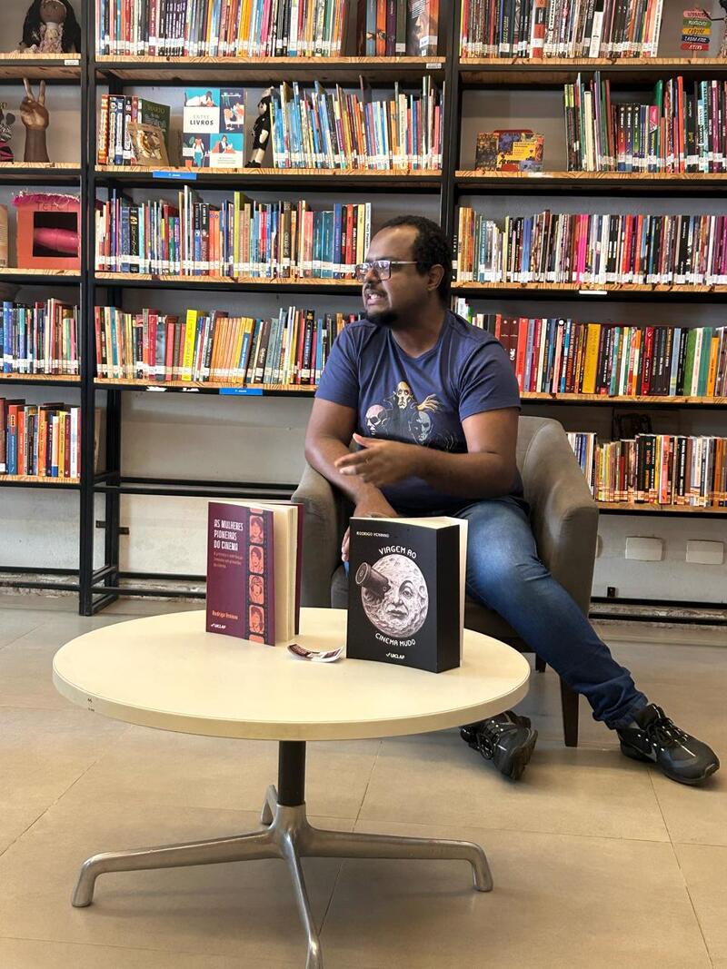 Imagem do lançamento do livro Viagem ao Cinema Mudo, de Rodrigo Veninno. O autor aparece sentado em uma poltrona e à frente de uma estante com livros. Um exemplar da obra está em uma mesa à frente dele.