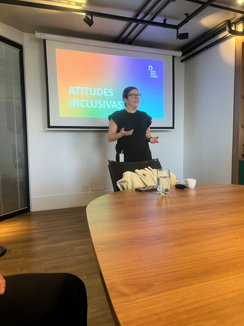 Facilitadora em pé apresentando slide sobre atitudes inclusivas diante de tela com cores lilás, azul-claro e laranja.