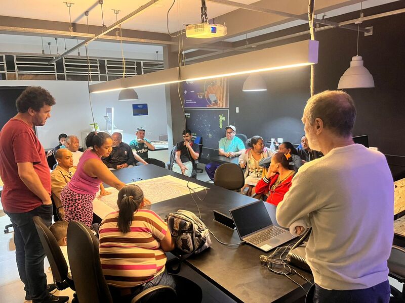 Imagem de pessoas reunidas no espaço coworking do Galpão ZL. Elas estão sentadas ao redor de uma mesa do espaço. Há também três pessoas em pé: um homem aparece de costas, à frente de um notebook, outro está mais para o canto esquerdo, de braços cruzados, e uma mulher aponta para uma folha A1 na qual aparece o mapa do Jardim Lapena. Há uma luminária sobre a mesa.