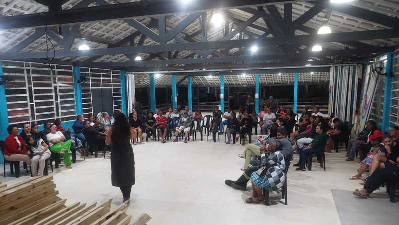 A imagem mostra uma reunião das pessoas integrantes do Plano de Bairro. Elas estão no auditório do Galpão ZL e aparecem sentadas em cadeiras distribuídas ao redor do espaço. Há uma pessoa no centro do pátio e em pé.