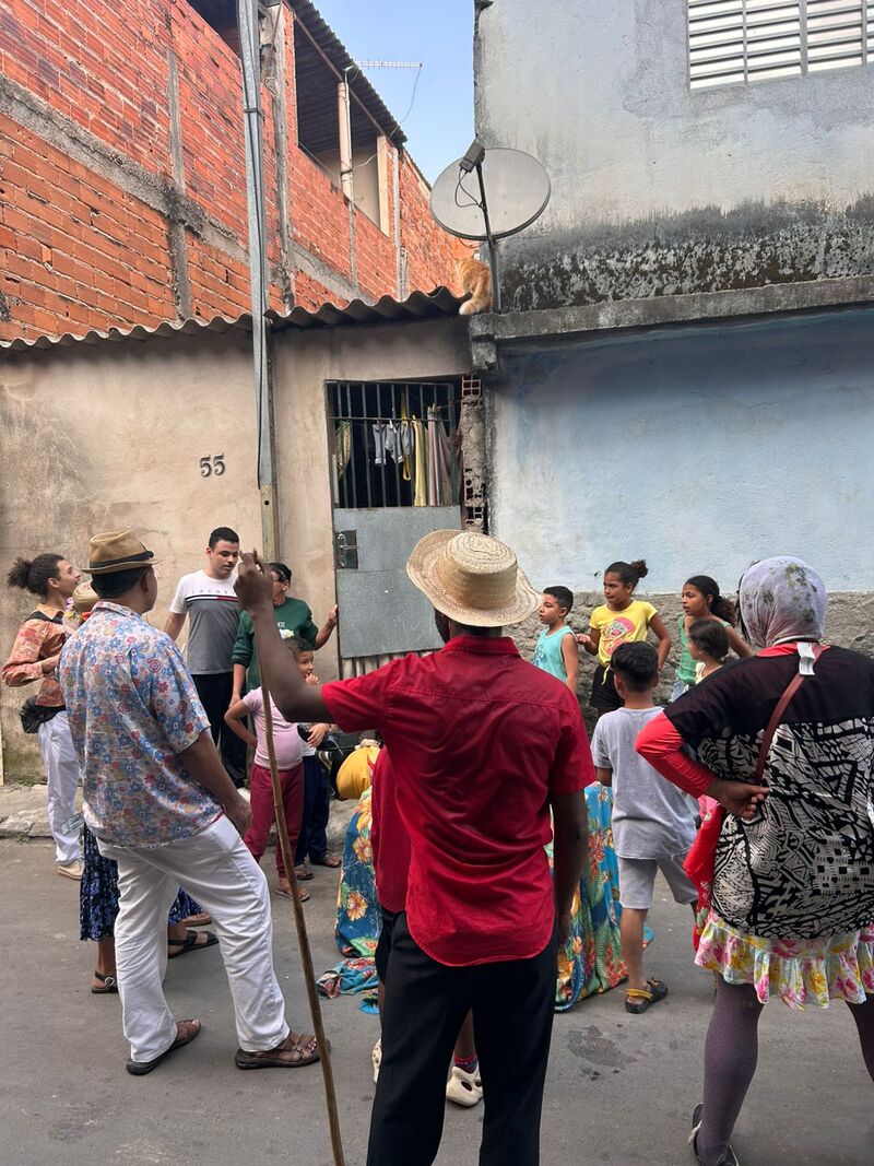 Imagem de uma atividade organizada pela equipe do Ponto de Leitura do Jardim Lapena. Pode-se ver um grupo de pessoas em uma viela do bairro. Um homem de camisa vermelha e chapéu de palha aparece apoiando-se em uma vara, enquanto crianças participam dessa mesma intervenção.